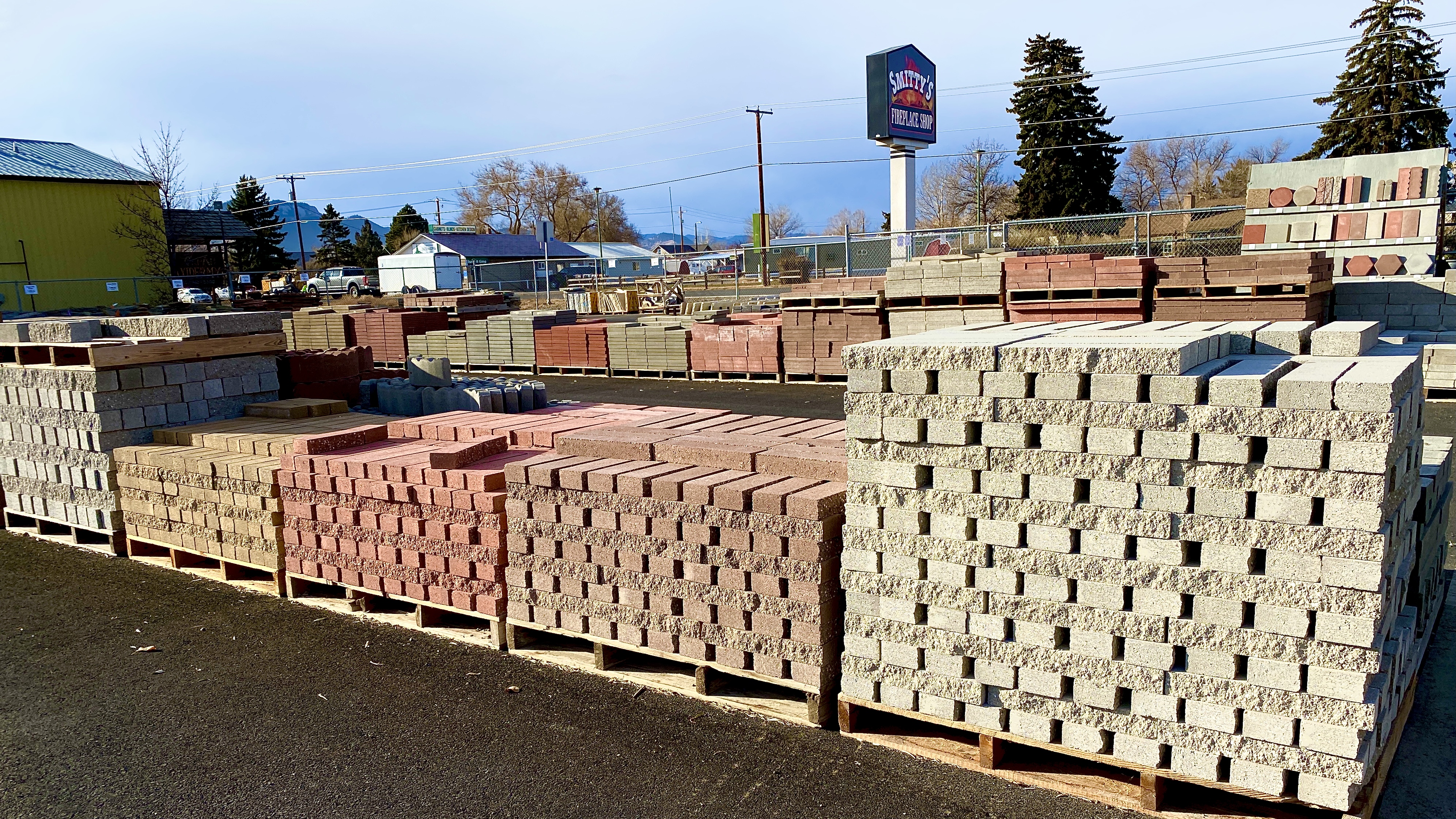 Stacks of masonry and landscaping cinder blocks stacked in a paved, fenced yard. Sign that says Smitty's Fireplace Shop in the background.